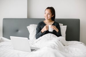 Young woman holding a coffee cup while sitting on her bed and a laptop in front of her.